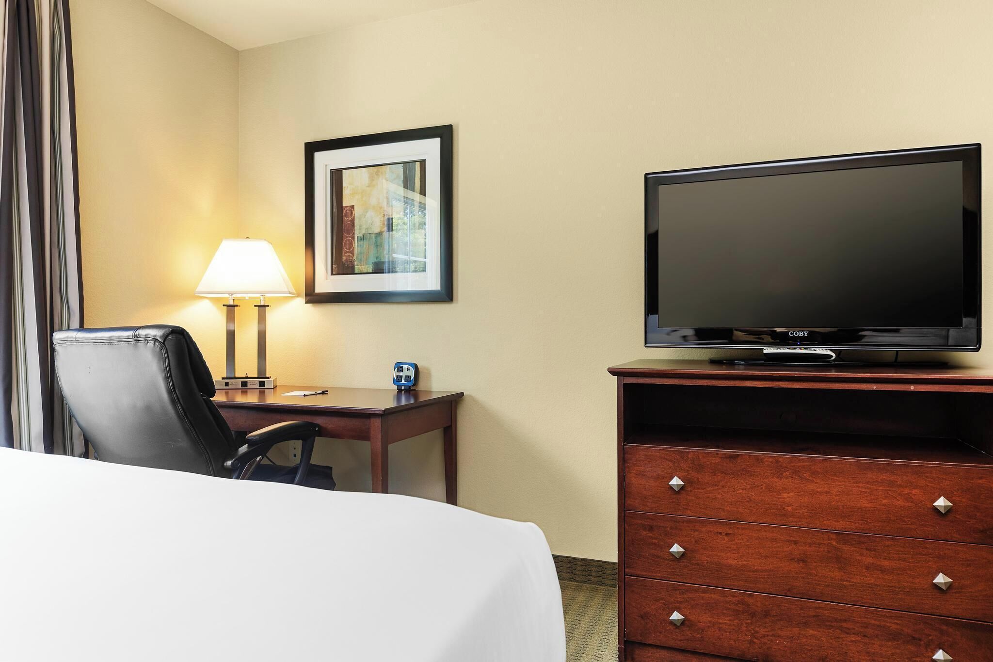 A hotel room with a bed, a desk with a lamp, a television on a wooden dresser, and a framed picture on the wall.