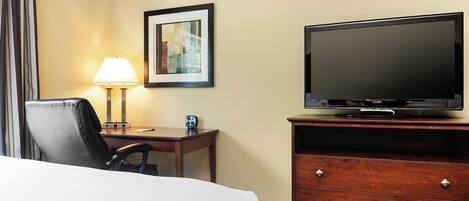 A hotel room with a bed, a desk with a lamp, a television on a wooden dresser, and a framed picture on the wall.