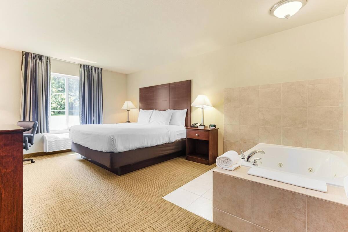 Une chambre d’hôtel comprenant un lit, des lampes de chevet, un bureau, une fenêtre avec des rideaux et une salle de bain avec une baignoire et un lavabo.