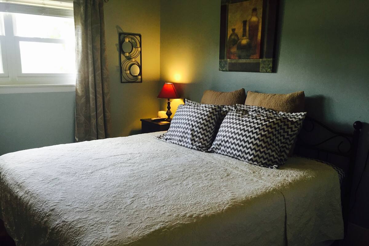Una cama bien hecha con un edredón blanco texturizado, dos almohadas estampadas, una lámpara de mesa roja y una obra enmarcada en la pared.