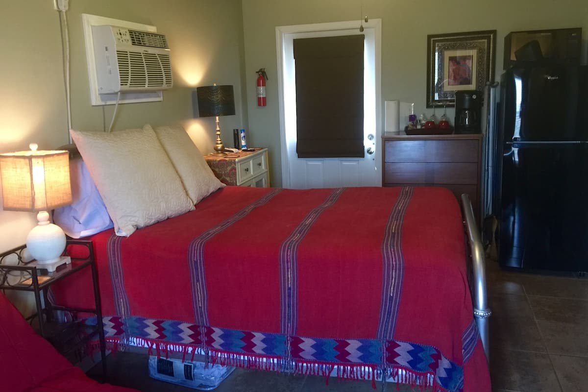 Una habitación de hotel con cama, mesita de noche, lámpara, refrigerador y una puerta.