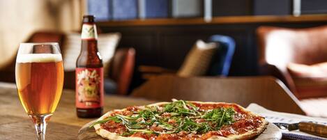 A pizza on a wooden board, a glass of beer, and a beer bottle on a table in a restaurant.