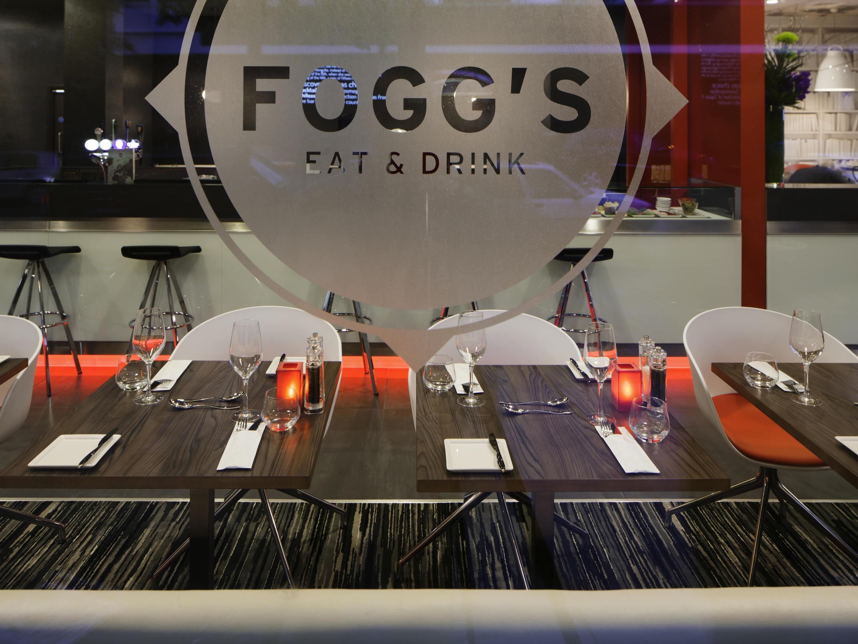 Interior de restaurante con mesas dispuestas para comer, una pared de vidrio con el nombre del restaurante y una zona de bar al fondo.