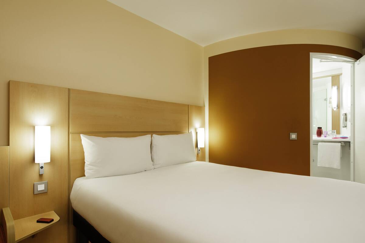 Habitación de hotel con una cama grande, lámparas de noche, mesita de noche y un baño visible a través de una puerta abierta.