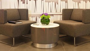 Um lobby moderno com dois poltronas cinzas, uma mesa de centro metálica e uma estante ao fundo.