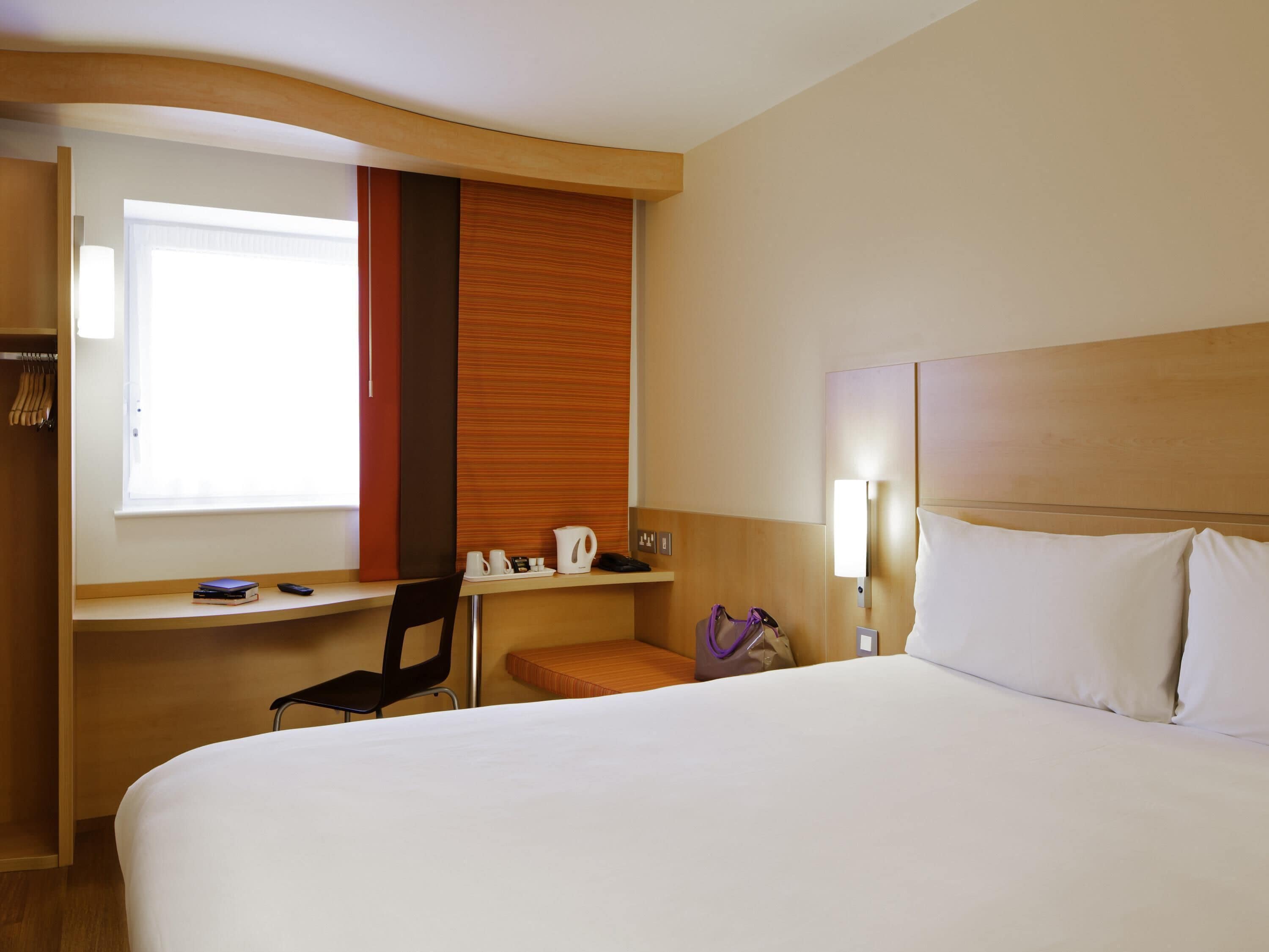 A hotel room with a bed, a desk with a chair, a window, and a wall-mounted light.