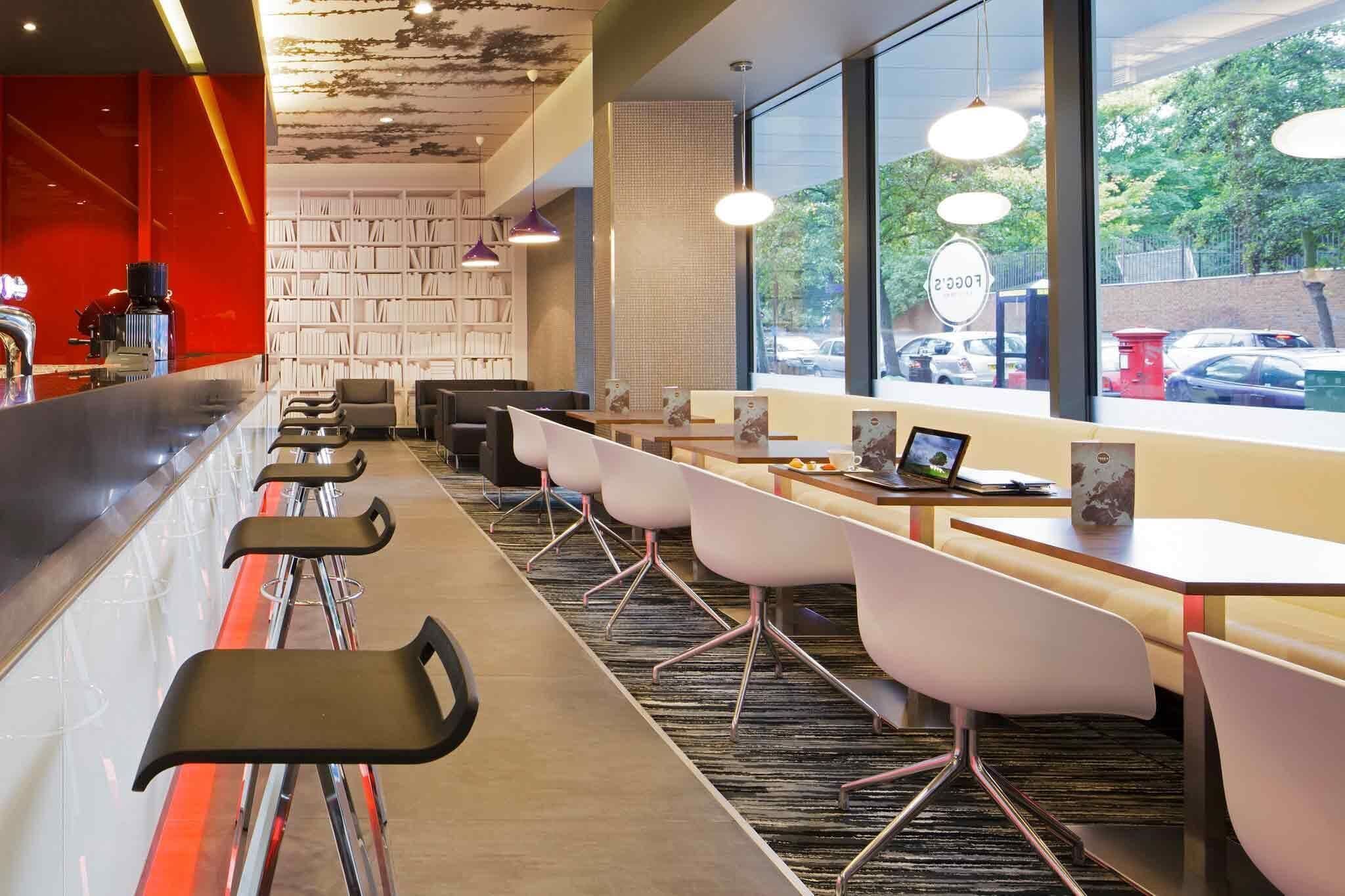 A modern cafe with a long counter, white chairs, and a large bookshelf.