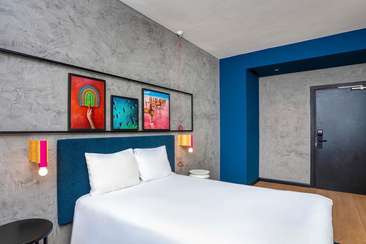 A modern hotel room with a large bed, artwork on the wall, a round table, and a door.