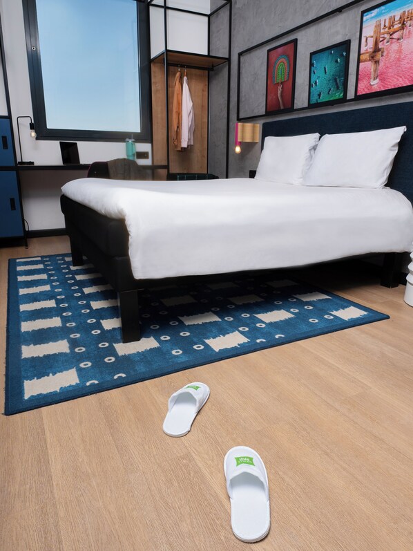 A modern hotel room with a bed, a blue patterned rug, a wooden floor, and a pair of slippers on the floor.