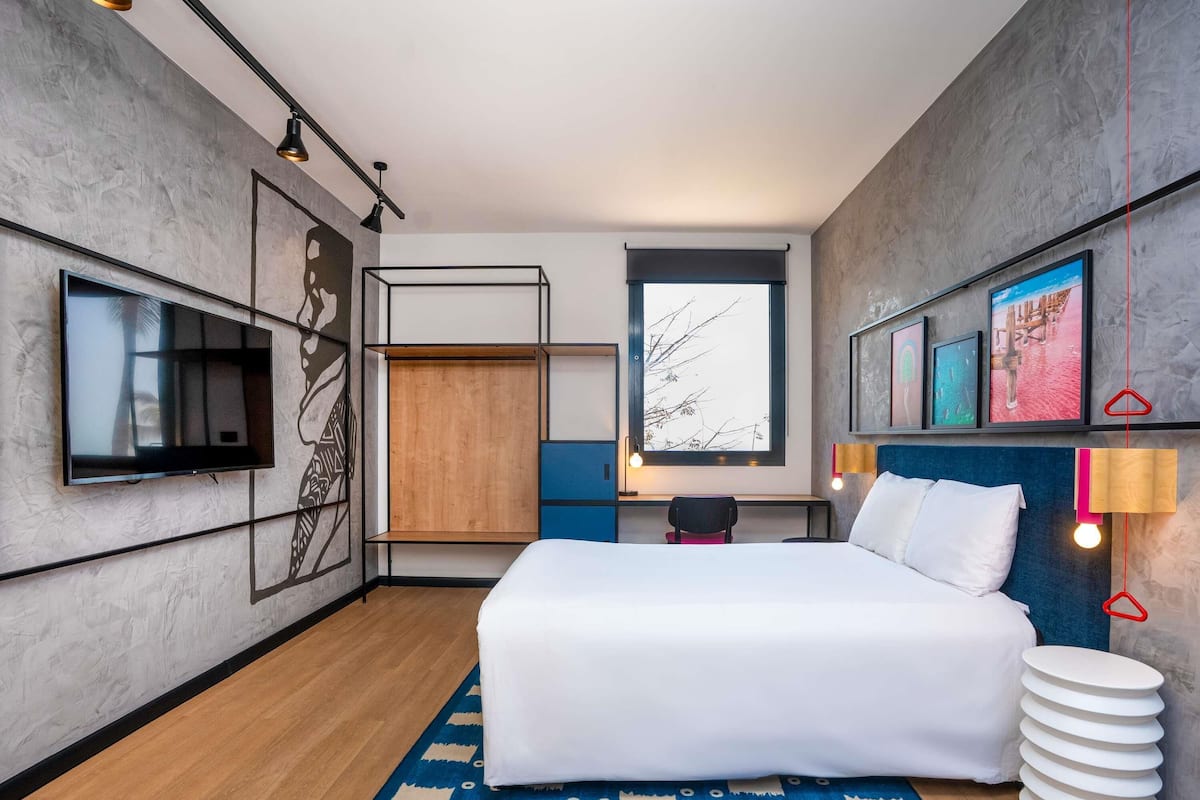 A modern bedroom with a bed, a TV, a desk, and a window with a view of trees.