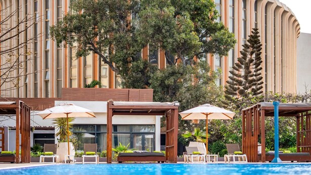 Una piscina con sillas de descanso y sombrillas, rodeada por un edificio de líneas verticales y un árbol grande.