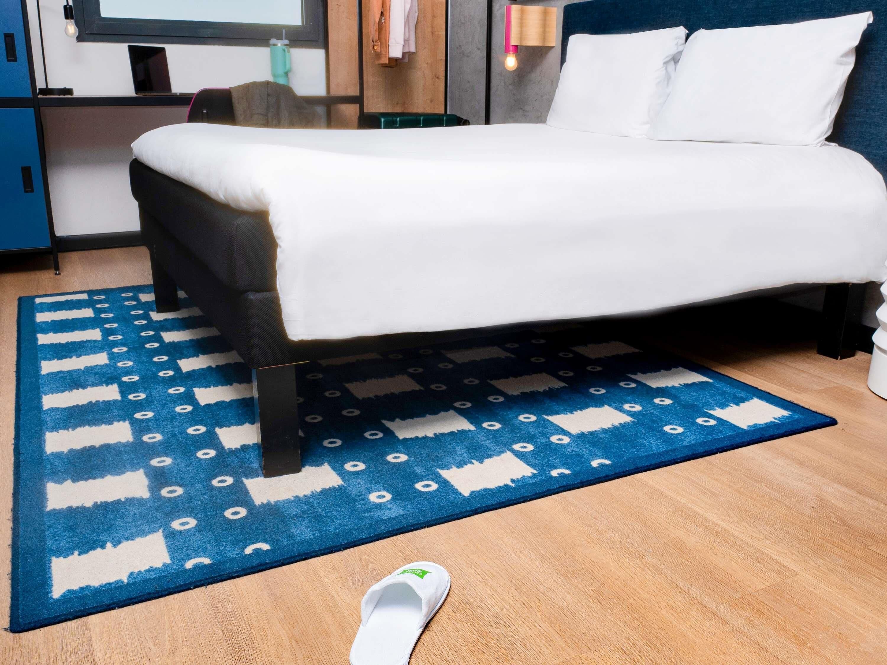 A neatly made bed with a white duvet, a blue patterned rug, and a single white slipper on the floor.