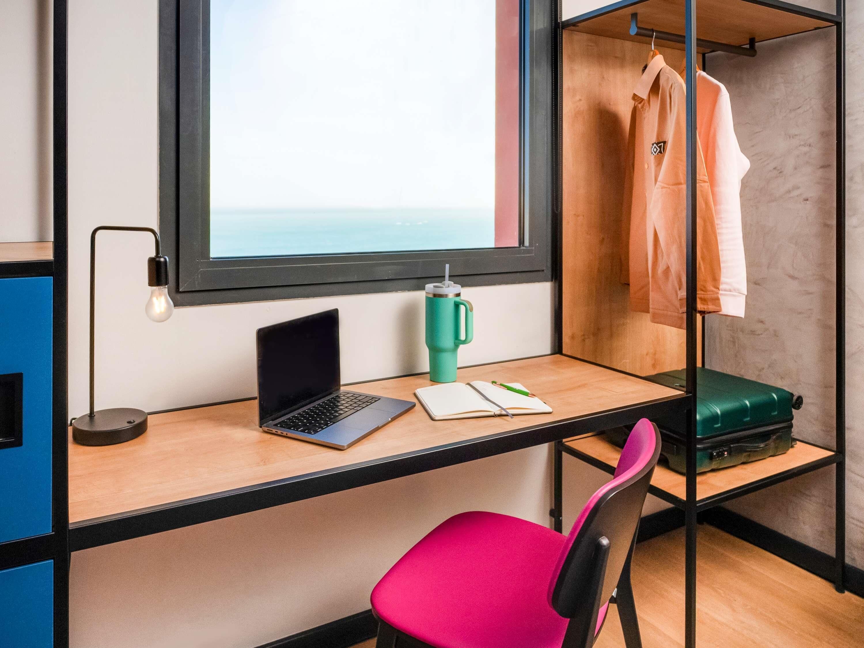A room with a desk, laptop, notebook, pen, green thermos, pink chair, and a suitcase.