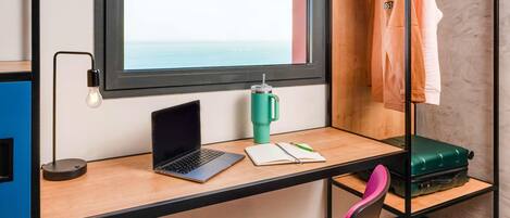 Una habitación con escritorio, computadora portátil, cuaderno, bolígrafo, termo verde, silla rosa y una maleta.
