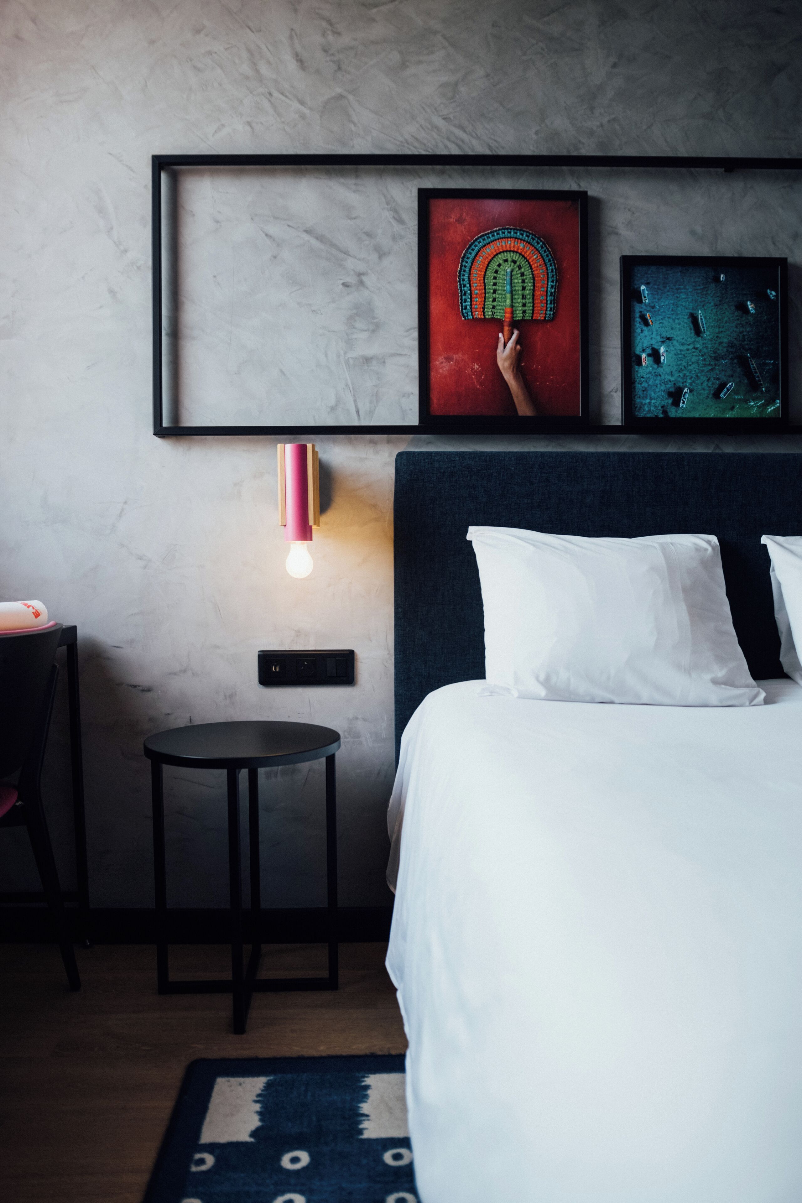 A hotel room with a bed, a small table, a wall-mounted light, and two framed artworks.