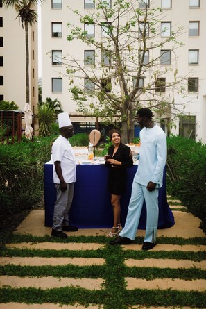 Eine Gruppe von Menschen steht an einem Buffet im Freien, mit einer blauen Tischdecke, umgeben von Pflanzen und einem Gebäude im Hintergrund.