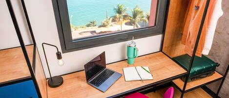 Une pièce avec un bureau, un ordinateur portable, un carnet, un stylo, une bouteille d’eau verte, une valise verte, une chaise rose et une fenêtre donnant sur l’océan et les palmiers.