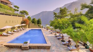 A pool area with lounge chairs and umbrellas, surrounded by greenery and mountains.