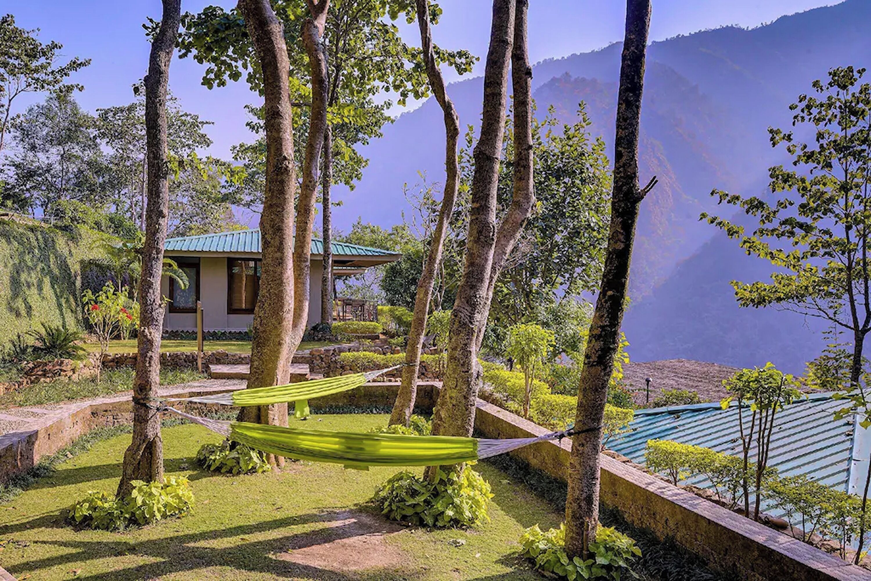 A serene garden with a hammock, a small building with a green roof, and a mountainous backdrop.