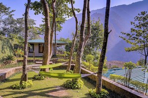 Un jardín tranquilo con una hamaca, un pequeño edificio con techo verde y un paisaje montañoso al fondo.