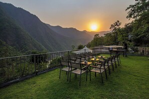 Un espacio de estar al aire libre con mesas y sillas, con vista a un paisaje montañoso durante el atardecer.