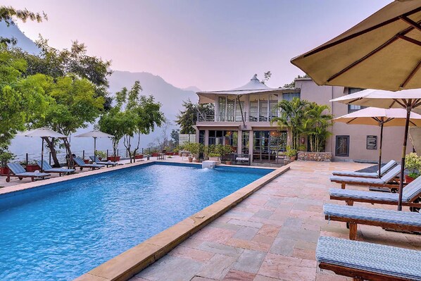 Piscina con sillas de descanso y sombrillas, rodeada por un edificio con vistas a montañas.