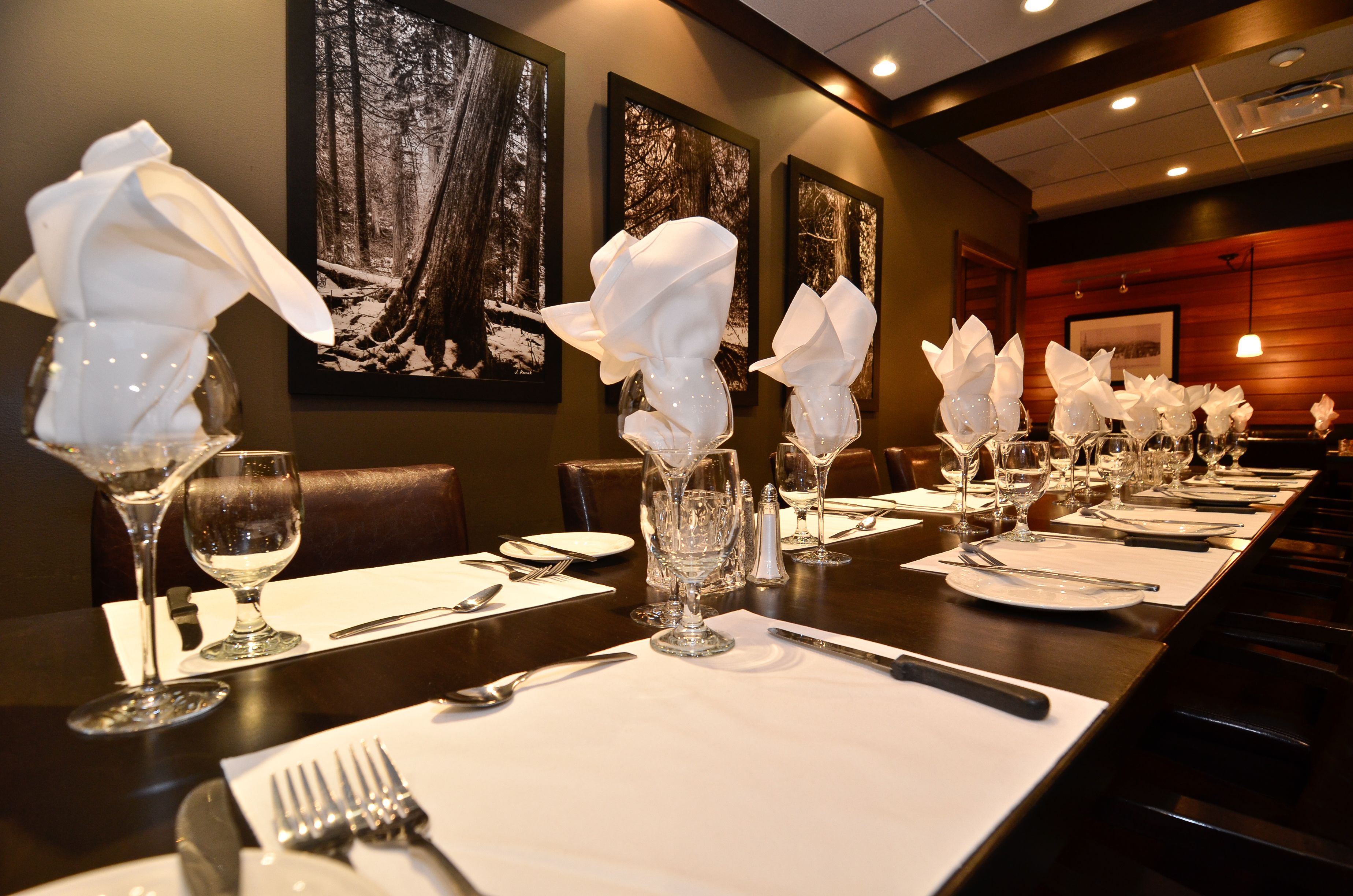 A dining table set with multiple wine glasses, plates, and cutlery, with a forest-themed wall art in the background.