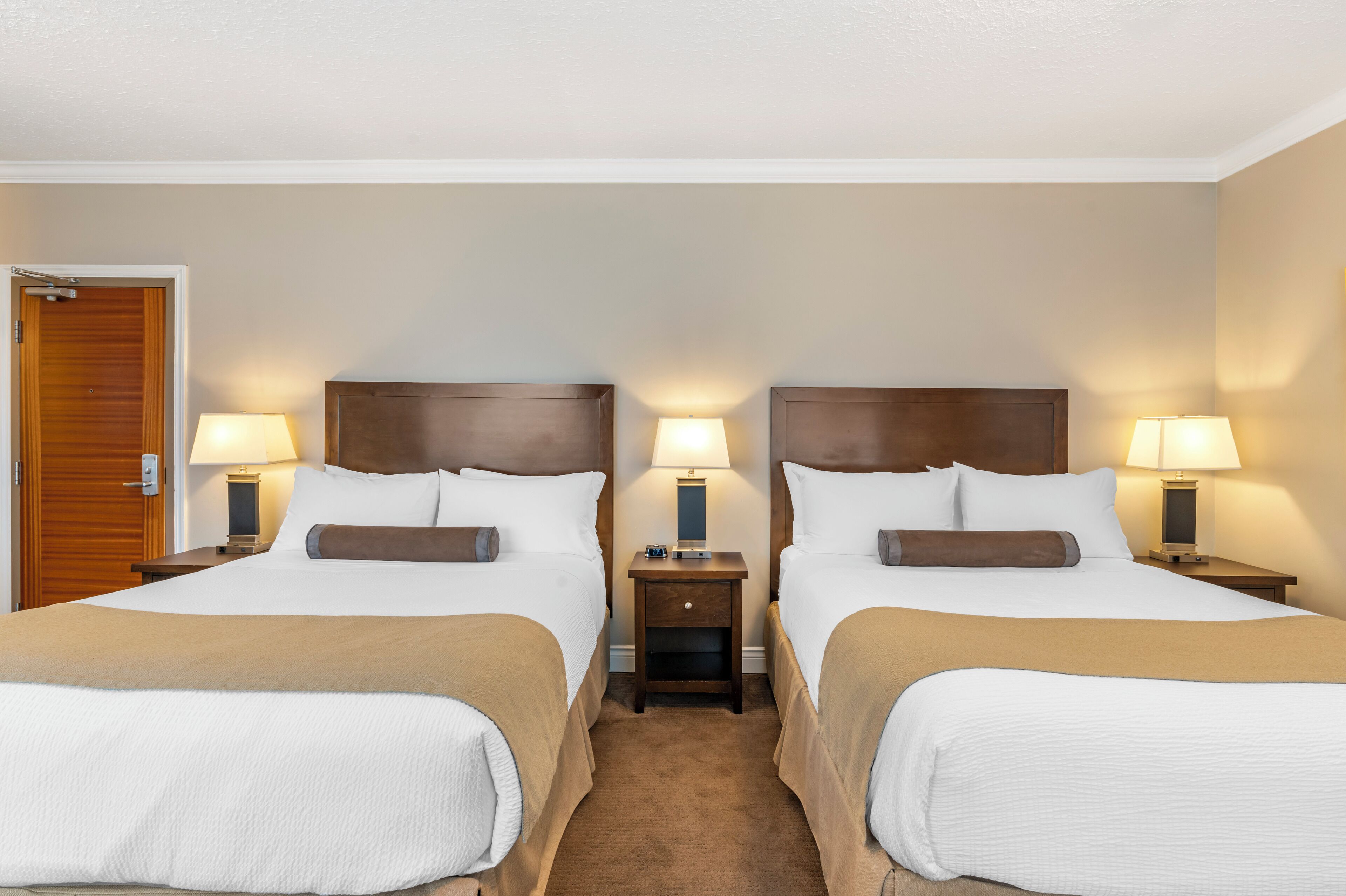 Quarto de hotel com duas camas, cada uma com cabeceira de madeira, mesa de cabeceira com abajur e uma porta à esquerda.