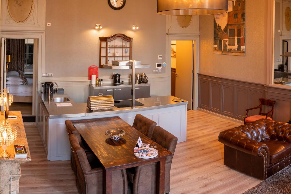Una cocina moderna con una isla central, un comedor con mesa de madera y sofá de cuero marrón, y un área de estar con un espejo grande y chimenea.