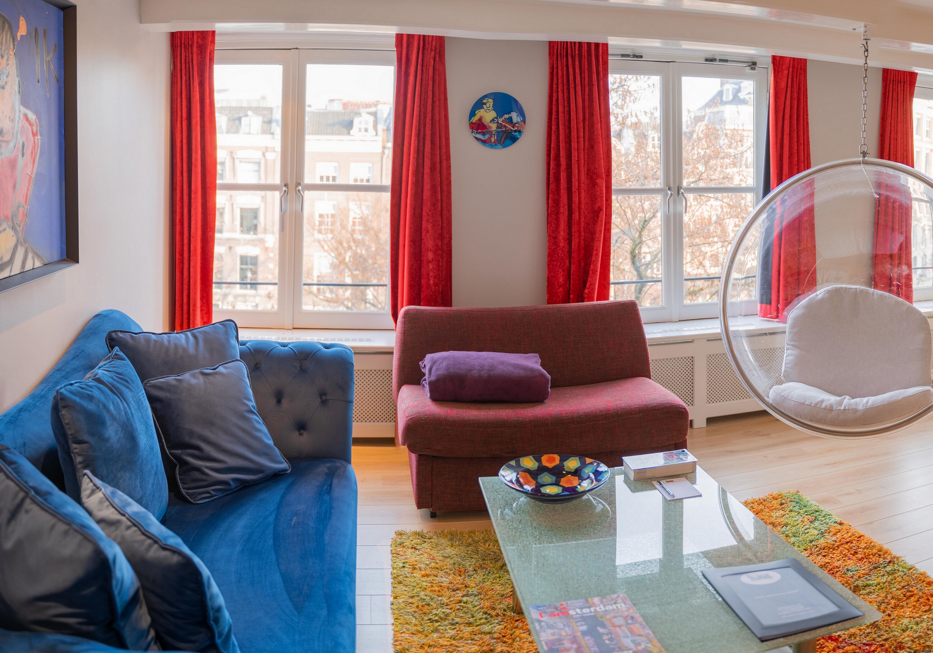 Una sala de estar con un sofá azul, un sillón rojo, una mesa de centro de vidrio y un asiento colgante.