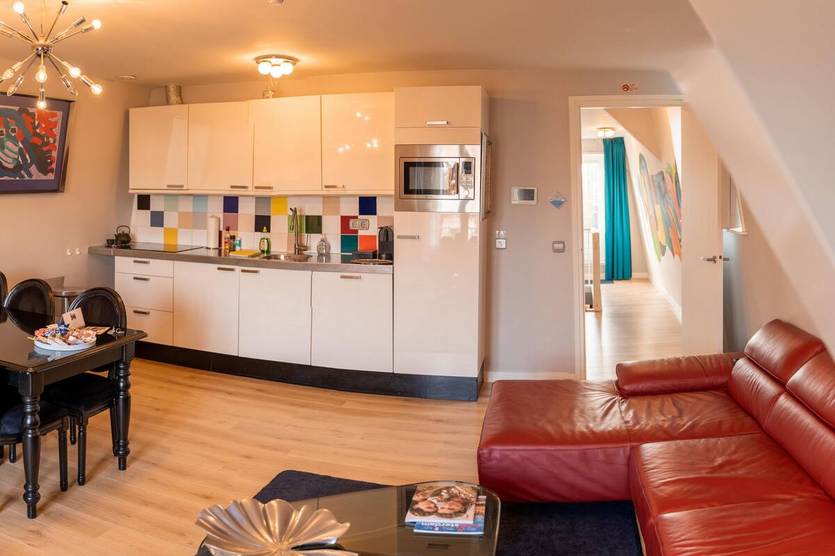 Cocina moderna con armarios blancos, un microondas y un salpicadero de colores. Hay un comedor con mesa y sillas, y un sofá de cuero rojo en la sala.