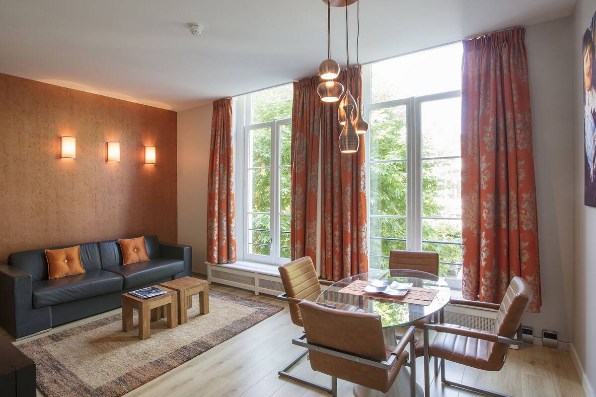 Una sala moderna con sofá, mesa de centro y mesa de comedor con sillas. Hay ventanas grandes con cortinas, una lámpara en el techo y un radiador.