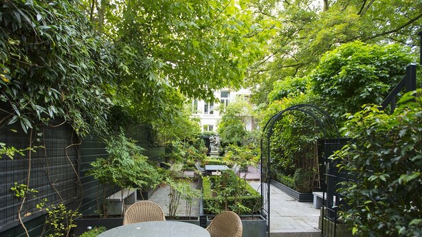A round table with wicker chairs in a garden area with a view of a building in the background. - Dutch Masters Amsterdam (Amsterdam)
