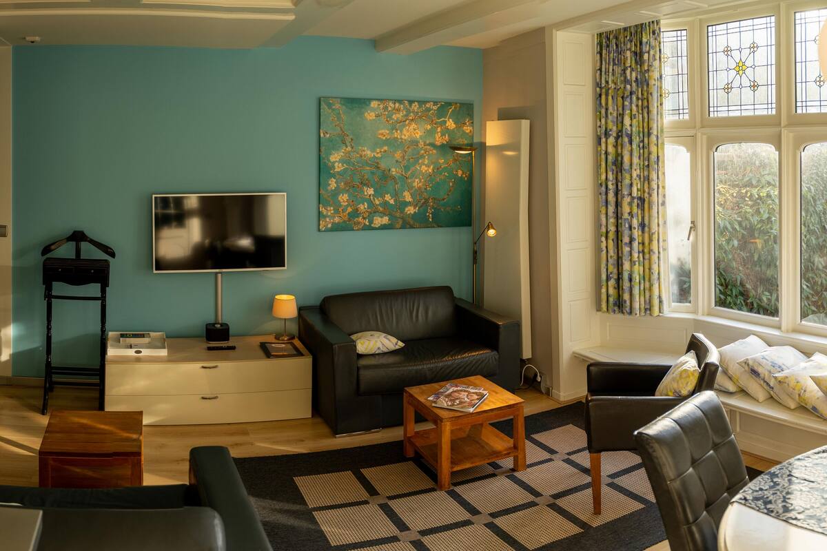 Un salón con pared verde azulado, un sofá de cuero negro, una mesa de centro de madera, un sillón negro, un televisor y una ventana con cortinas.