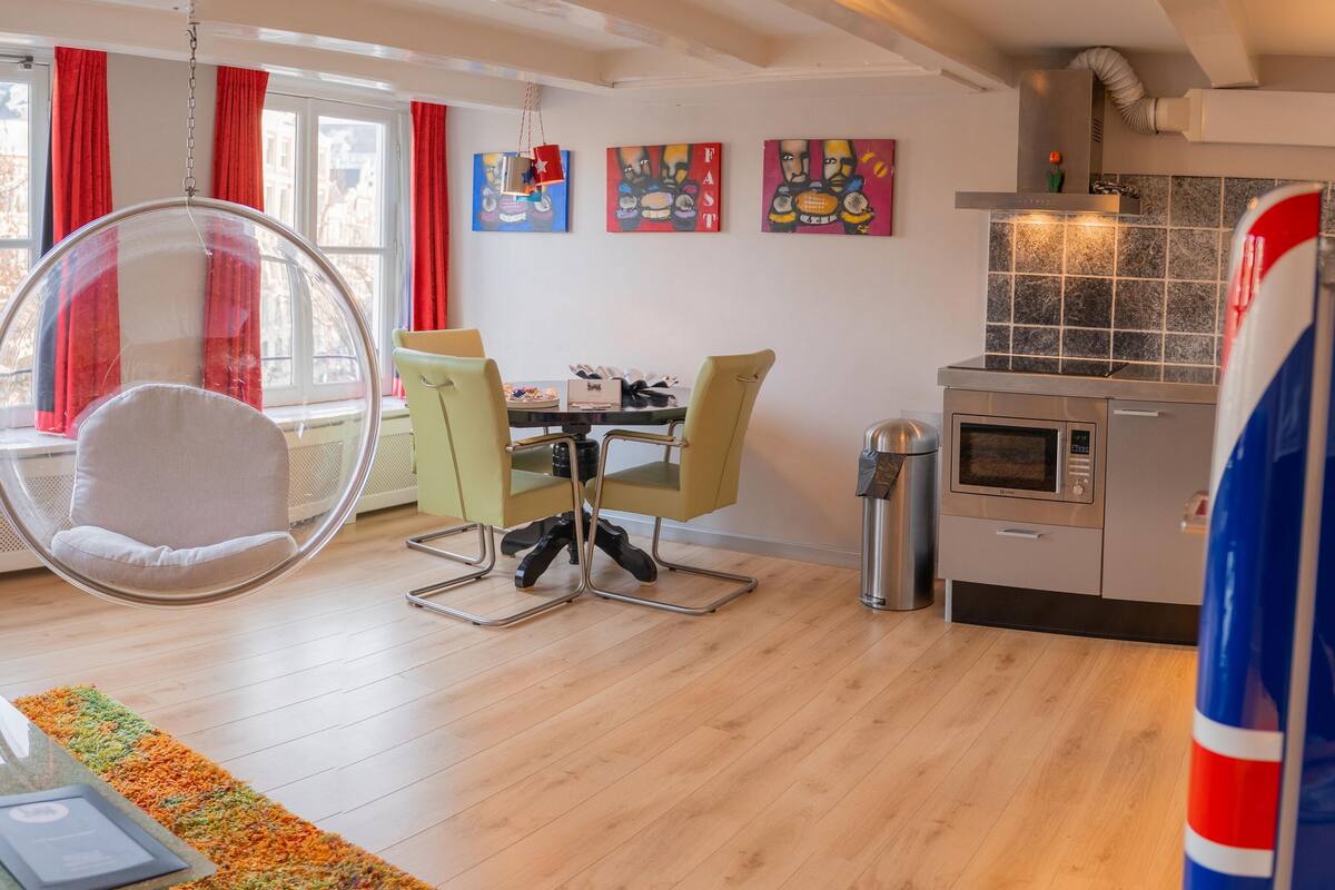 Una sala de estar moderna con una silla colgante, una mesa de comedor con sillas y una zona de cocina con microondas y un cubo de basura.