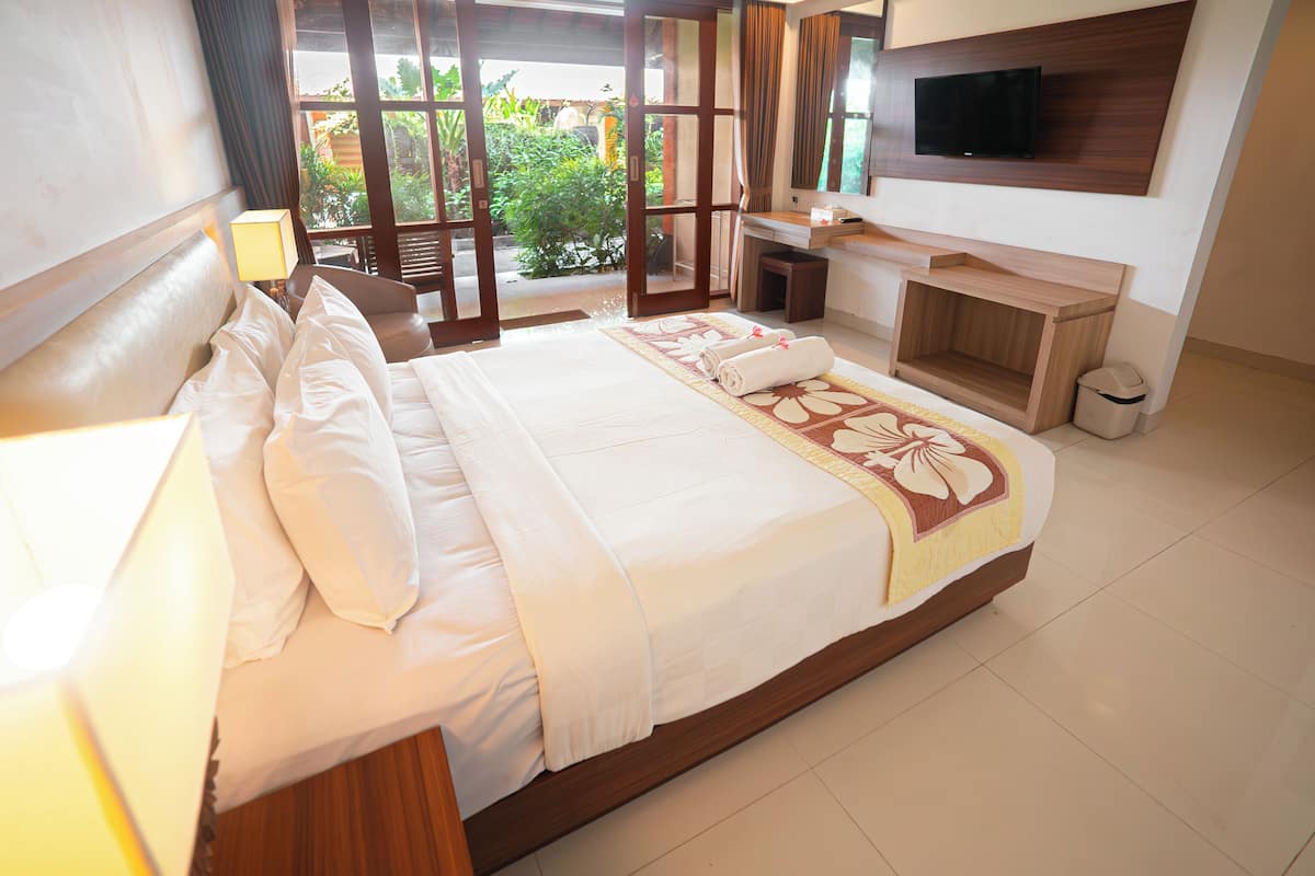 A hotel room with a large bed, a TV mounted on the wall, a bathroom with a sink and mirror, and a view of greenery outside.