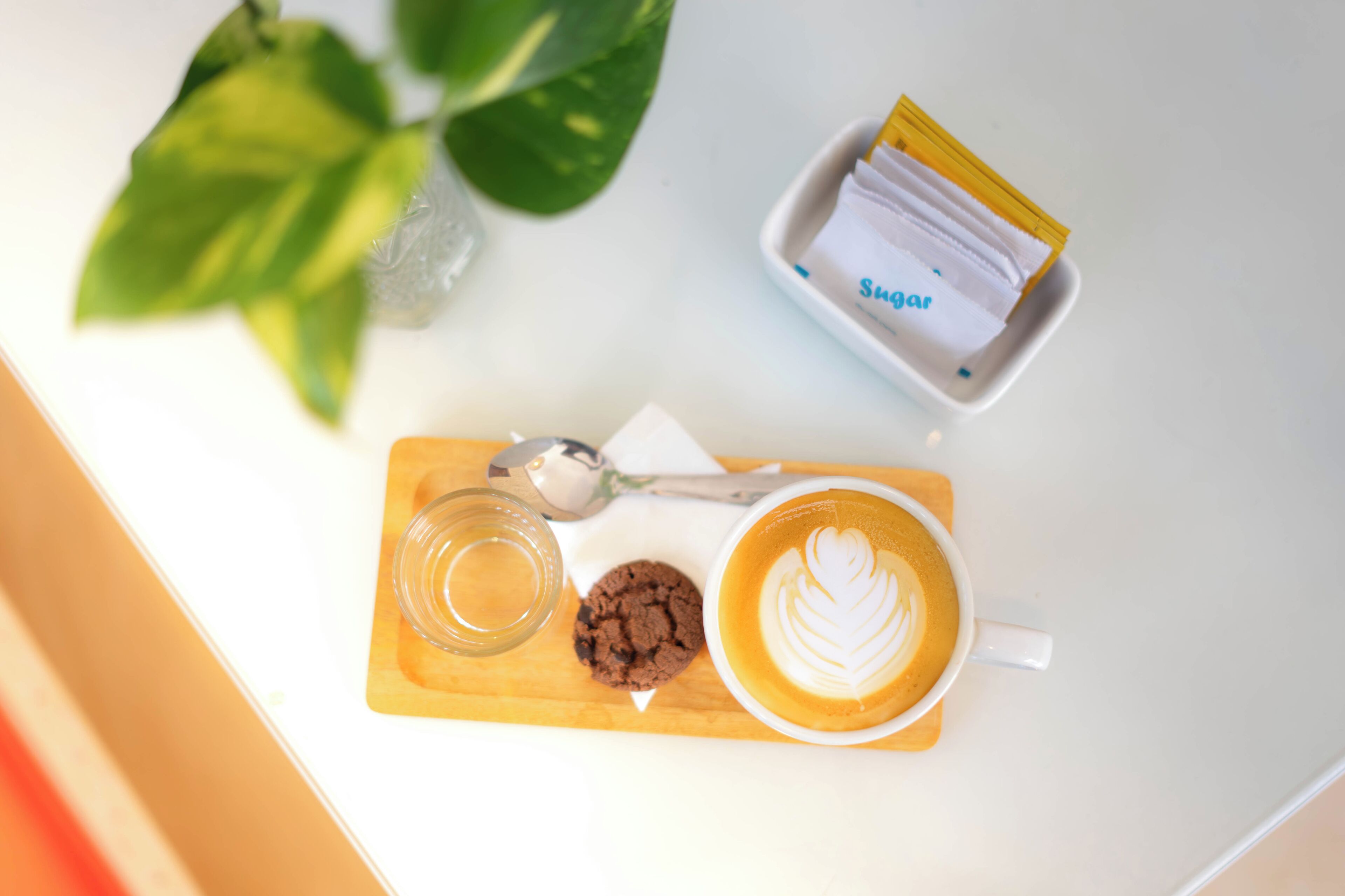 A coffee cup with latte art, a cookie, a glass of water, and a sugar packet on a wooden board.
