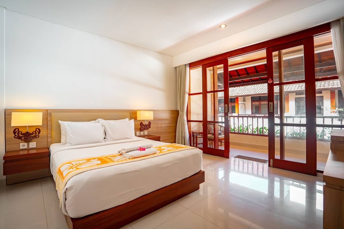 A hotel room with a large bed, wooden headboard, two bedside tables with lamps, and a view of a traditional building through a glass door.