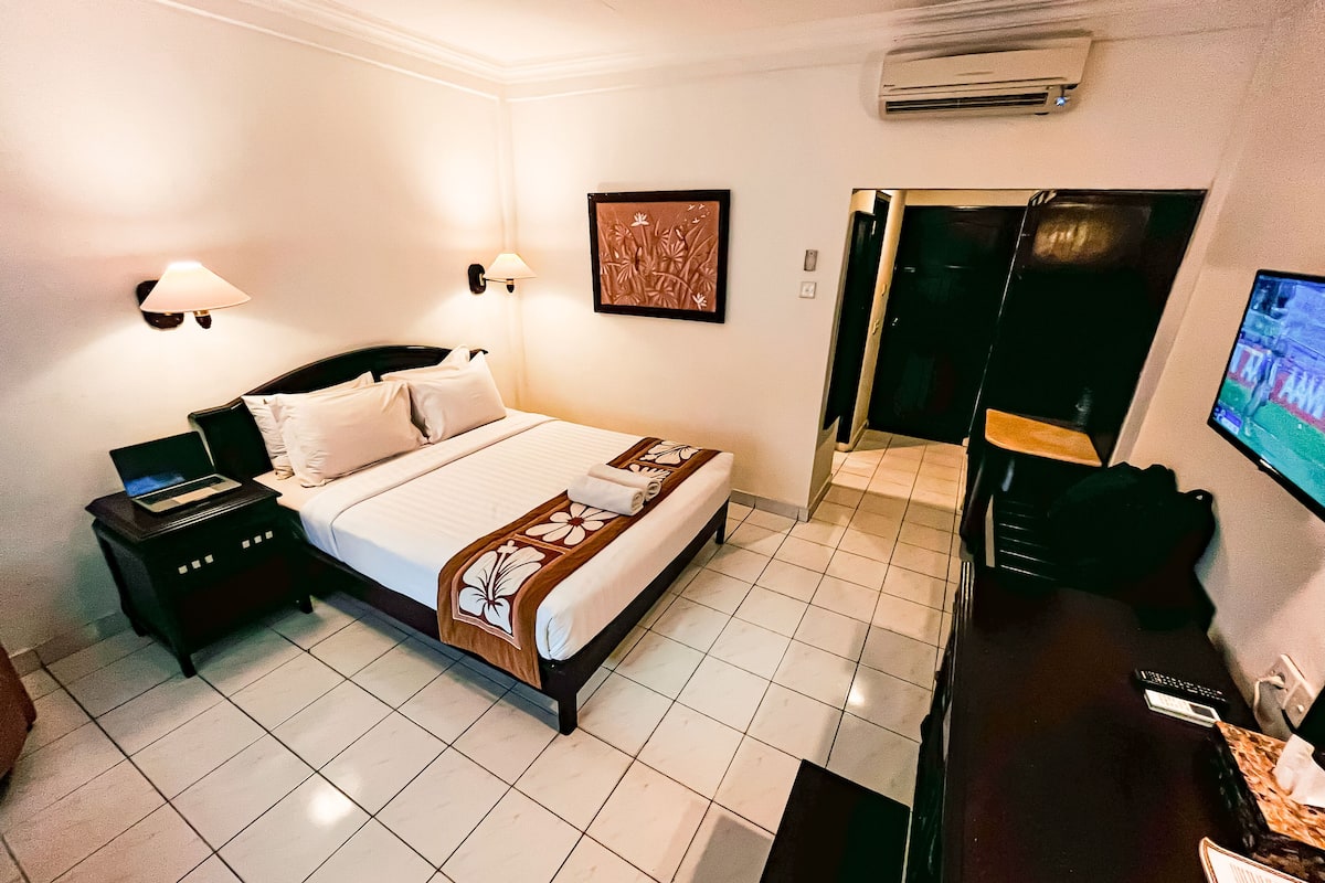 A hotel room with a bed, bedside tables, a desk with a computer, a TV, and an air conditioning unit.