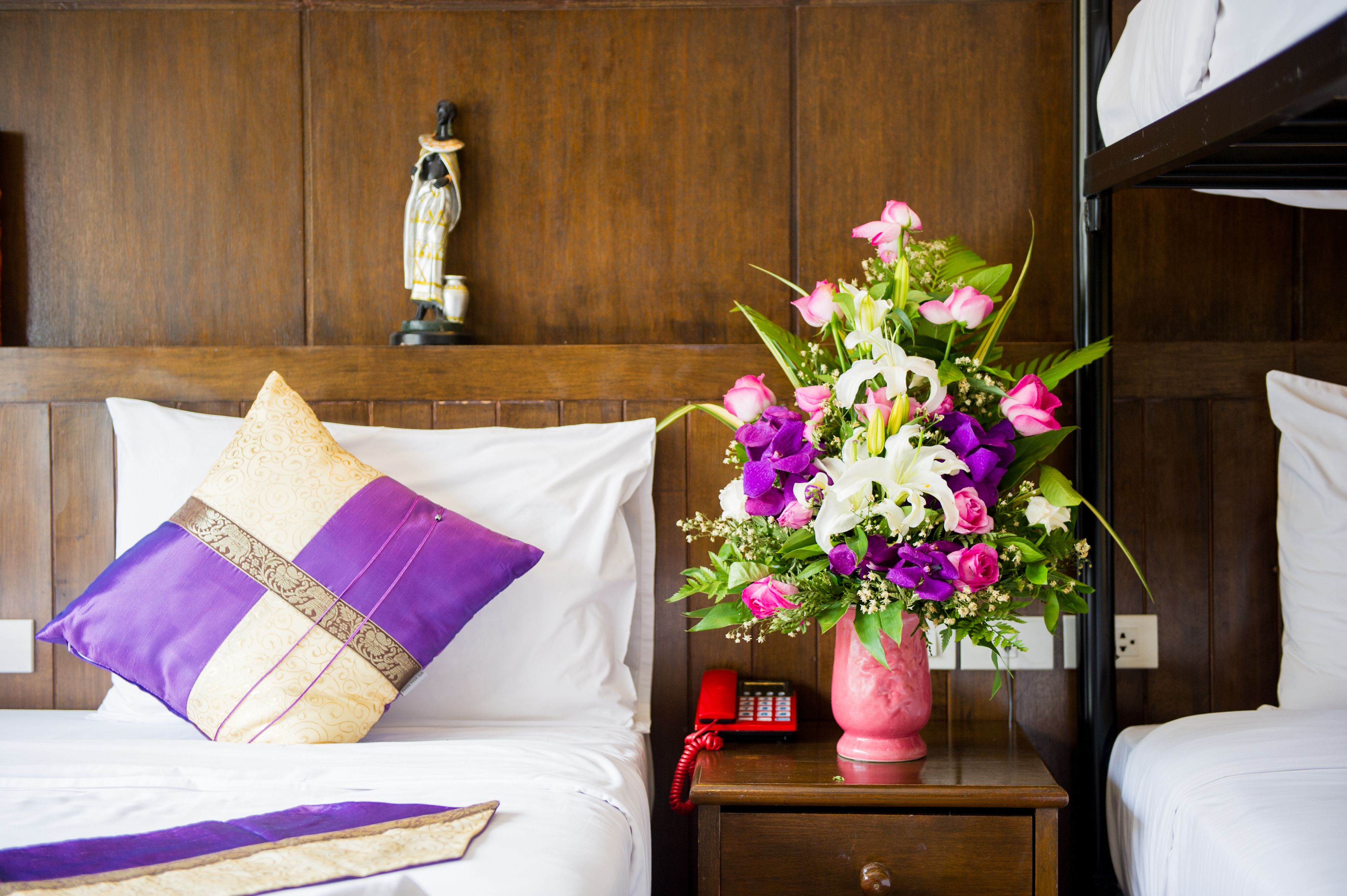 A hotel room with a wooden headboard, a bed with white linens and a purple and gold pillow, a nightstand with a red telephone, and a vase of flowers.