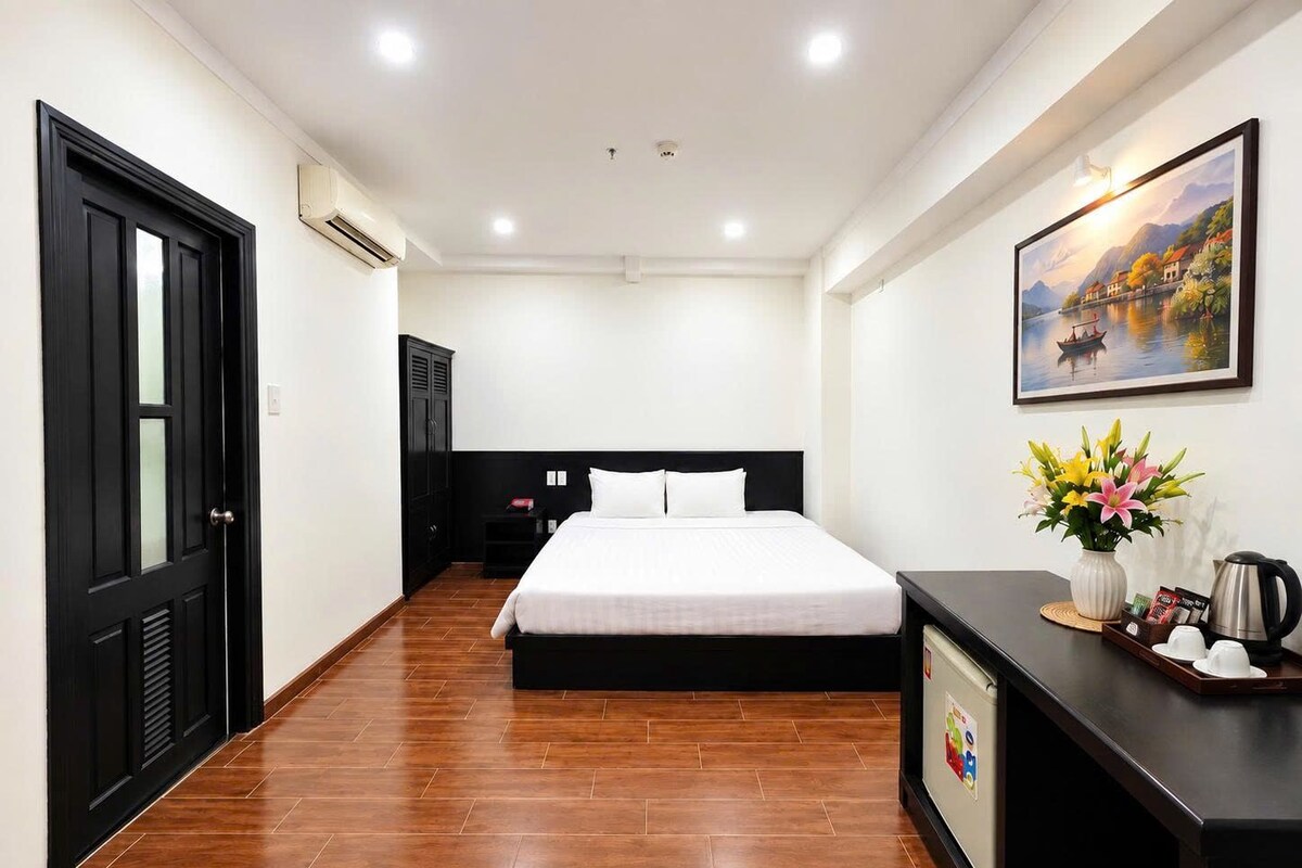 A hotel room with a bed, a desk with a vase of flowers, a painting on the wall, and a door.
