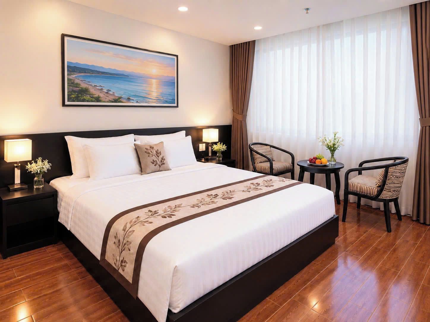 A hotel room with a large bed, two bedside tables with lamps, a small round table with fruit and flowers, and a view of the sea through sheer curtains.