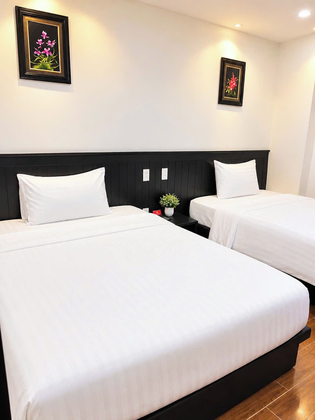 Ein Hotelzimmer mit zwei Betten, einem schwarzen Kopfteil, weißer Bettwäsche und zwei gerahmten Blumenbildern an der Wand.