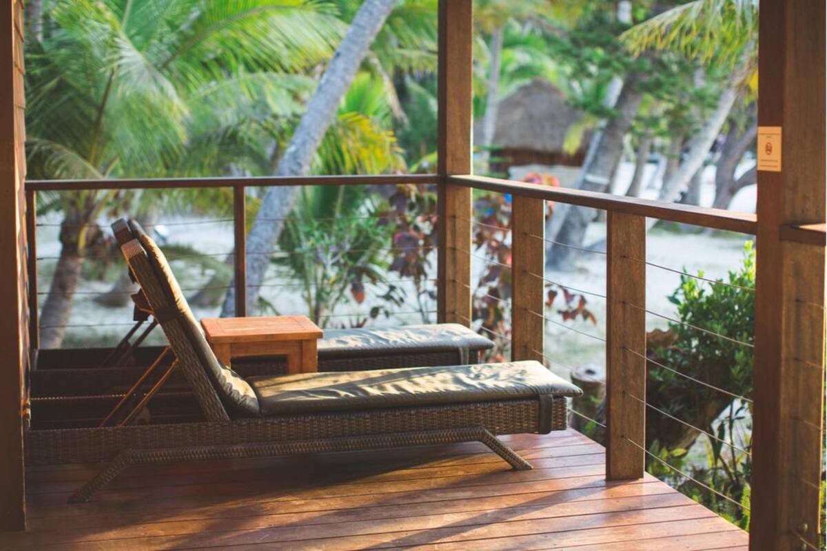 Une terrasse en bois avec des fauteuils de jardin en osier et une petite table, donnant sur une plage bordée de palmiers.