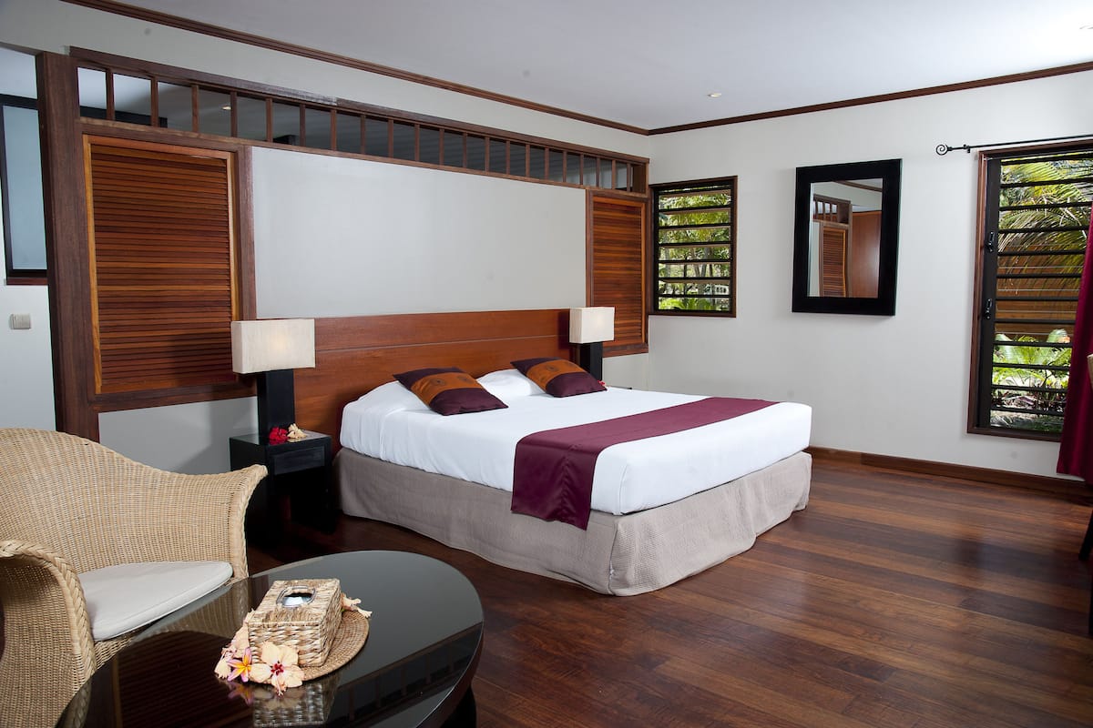 Une chambre avec une tête de lit en bois, un grand lit avec un matelas blanc et une couverture bordeaux, une chaise en osier, une petite table avec un panier, et une fenêtre donnant sur un paysage verdoyant.