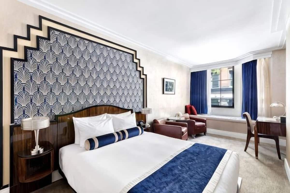 A hotel room with a large bed, a desk, a chair, a nightstand, and a window with curtains.