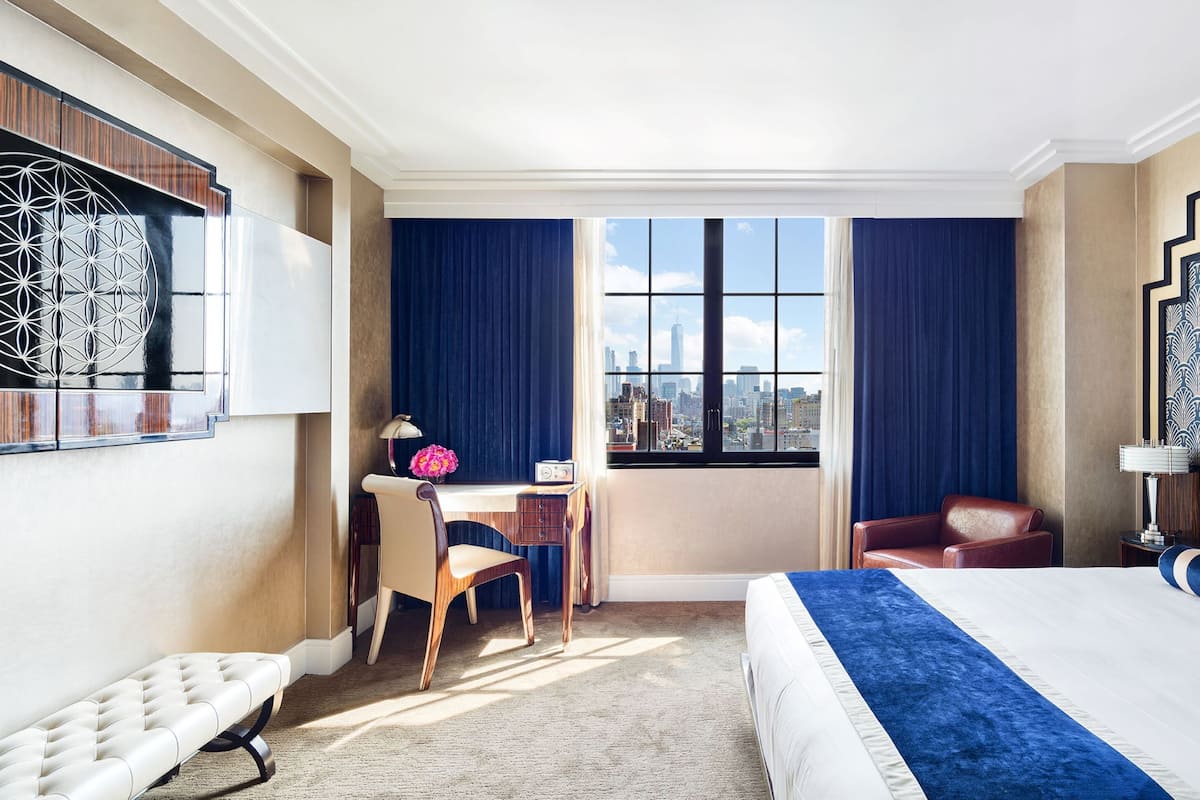 A hotel room with a large bed, a desk with a chair, a window with a city view, and a patterned headboard.
