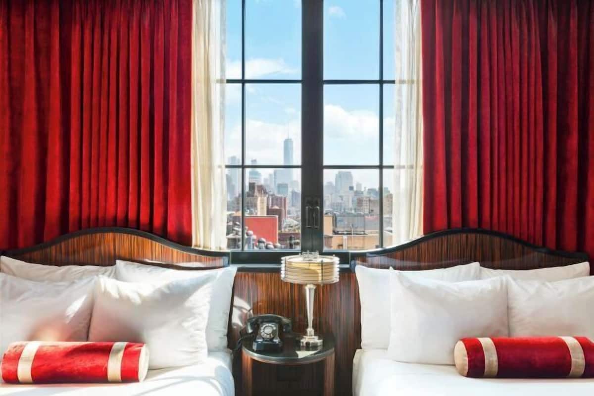 A hotel room with a large window, two beds, a nightstand with a lamp, and a city view.
