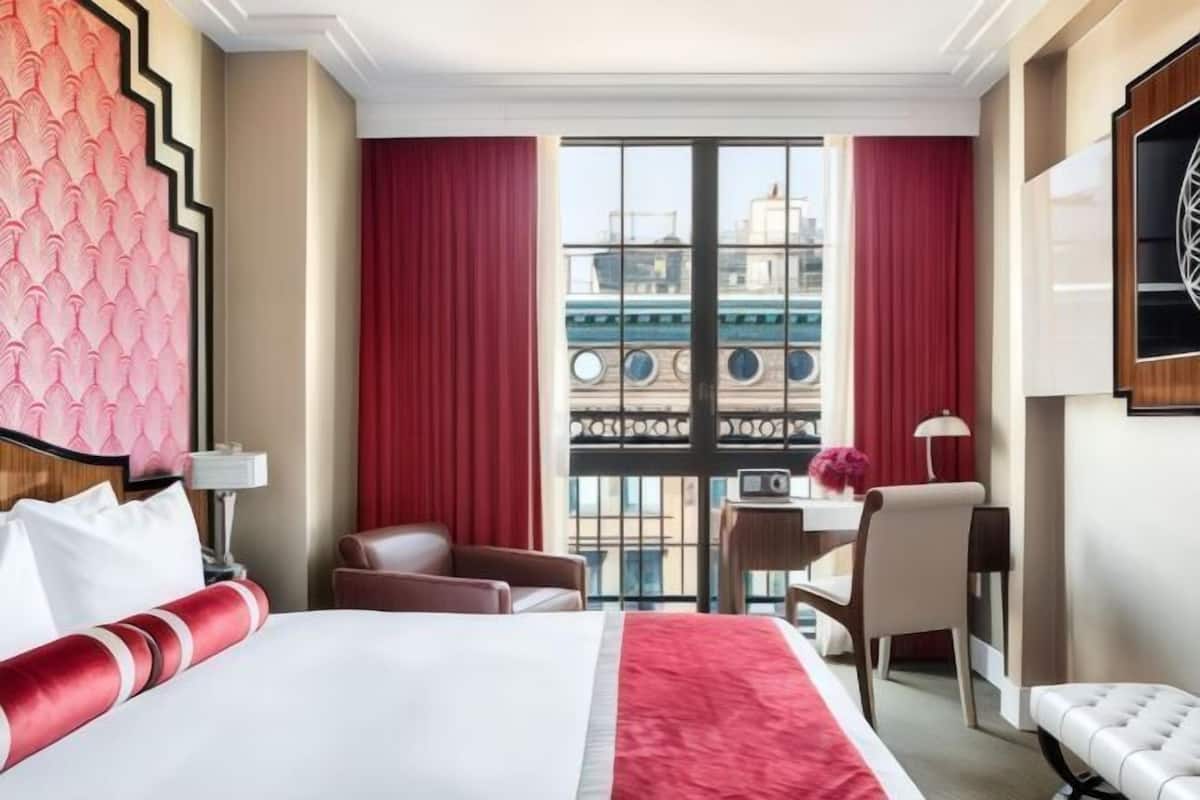 A hotel room with a large bed, a desk, a chair, and a view of buildings through a window.