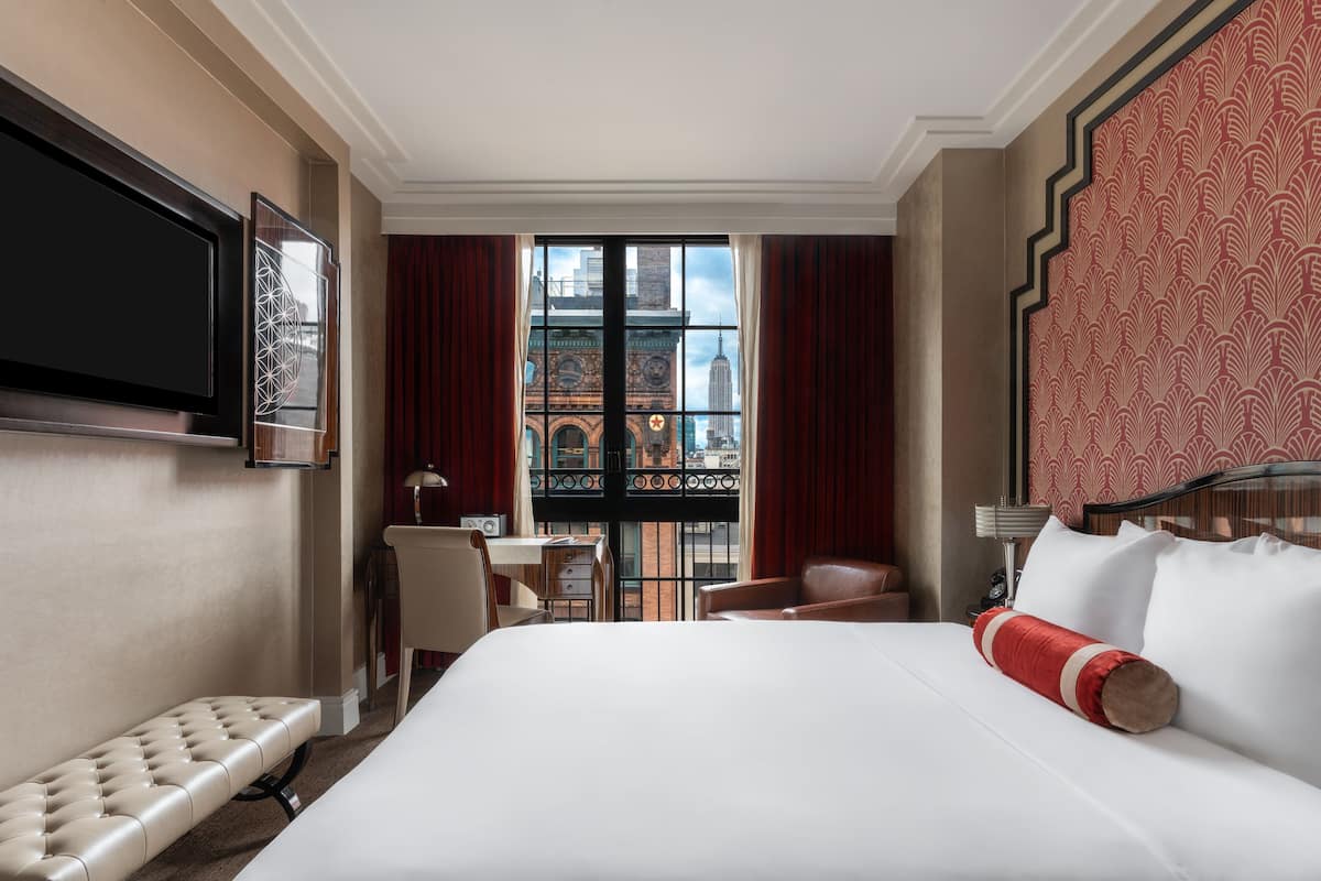 A hotel room with a large bed, a desk, and a chair. There is a TV mounted on the wall and a window with a view of buildings.